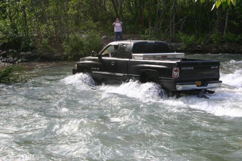 2003 Jeep TJ - A black Dodge Ram pickup truck navigates through a shallow river, kicking up water.
