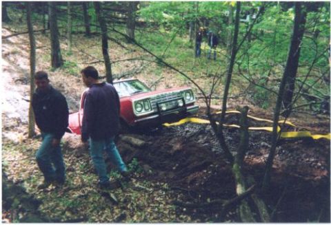 1996 Ford Bronco - A red full-size SUV with a white roof and stacked square headlights is nosed down into a muddy wooded trail rut, front bumper buried and a…
