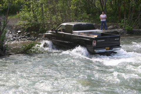 2003 Jeep TJ - A black Dodge Ram pickup truck navigates through a shallow river with trees and brush on the banks.