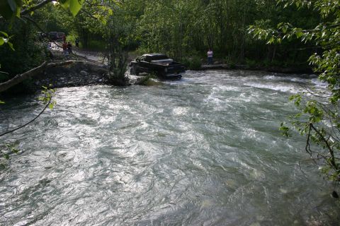 2003 Jeep TJ - A black pickup truck navigates a shallow river with trees lining the banks and a few people observing.