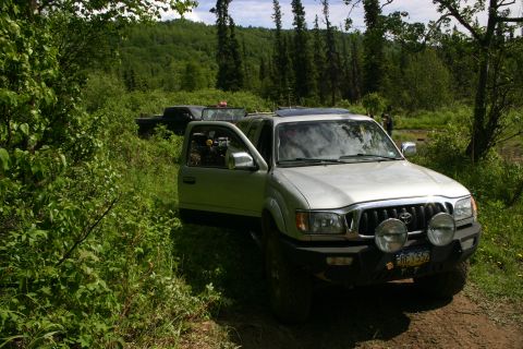2003 Jeep TJ - A silver 2003 Toyota Tacoma pickup truck is parked off-road with its driver door open, alongside another dark-colored truck in the backgrou…