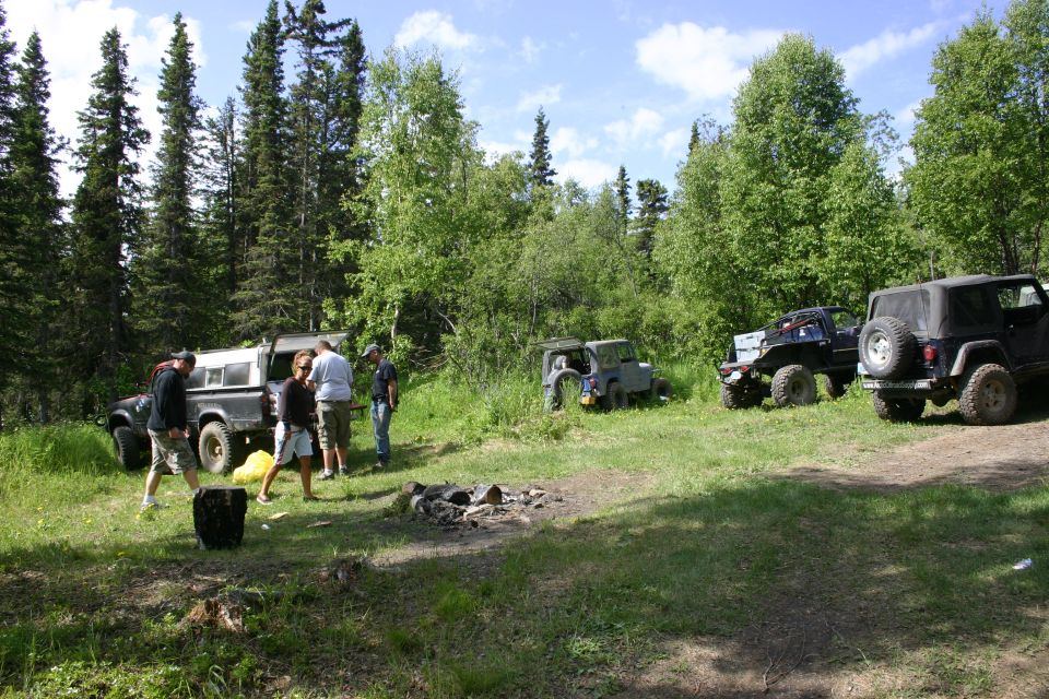 Several off-road vehicles parked in a grassy clearing surrounded by trees, with people standing near one of them