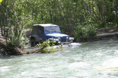 2003 Jeep TJ - A blue Jeep Wrangler navigates a shallow river surrounded by dense foliage.