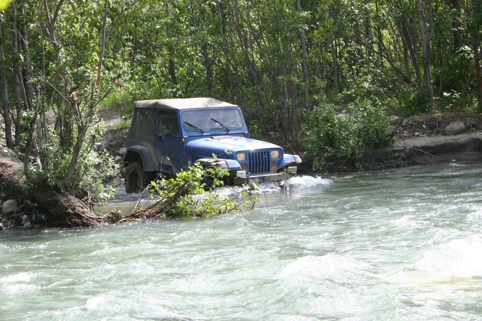 Blue Jeep Wrangler driving through a river with trees on both sides