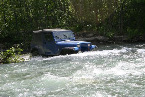 2003 Jeep TJ - A blue Jeep Wrangler navigates a rocky river, with trees lining the bank.