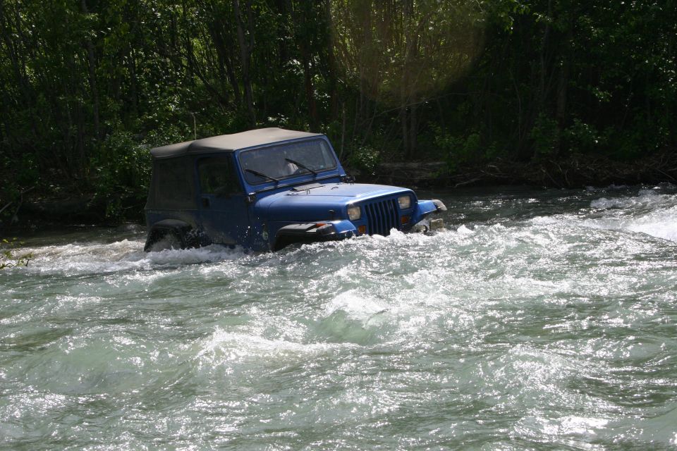 Blue 2003 Jeep TJ driving through a fast-moving river with trees in the background