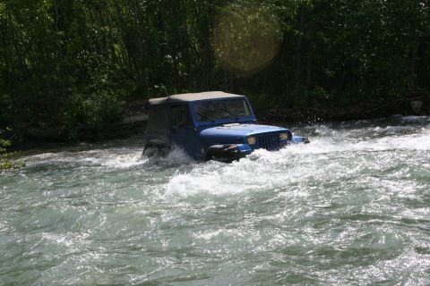2003 Jeep TJ - A blue Jeep Wrangler navigates a shallow river with trees lining the banks.