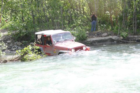 2003 Jeep TJ - A red 2003 Jeep TJ navigates a shallow river surrounded by trees, with a bystander observing from the bank.