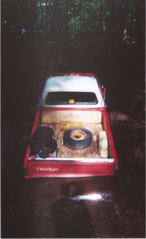 Looking down into red Dodge pickup bed with spare tire on wood floor, muddy forest trail at dusk