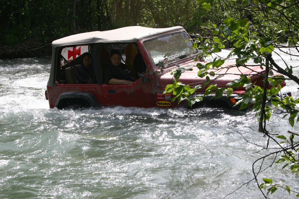 red Jeep Wrangler with a white soft top driving through a river, two people inside