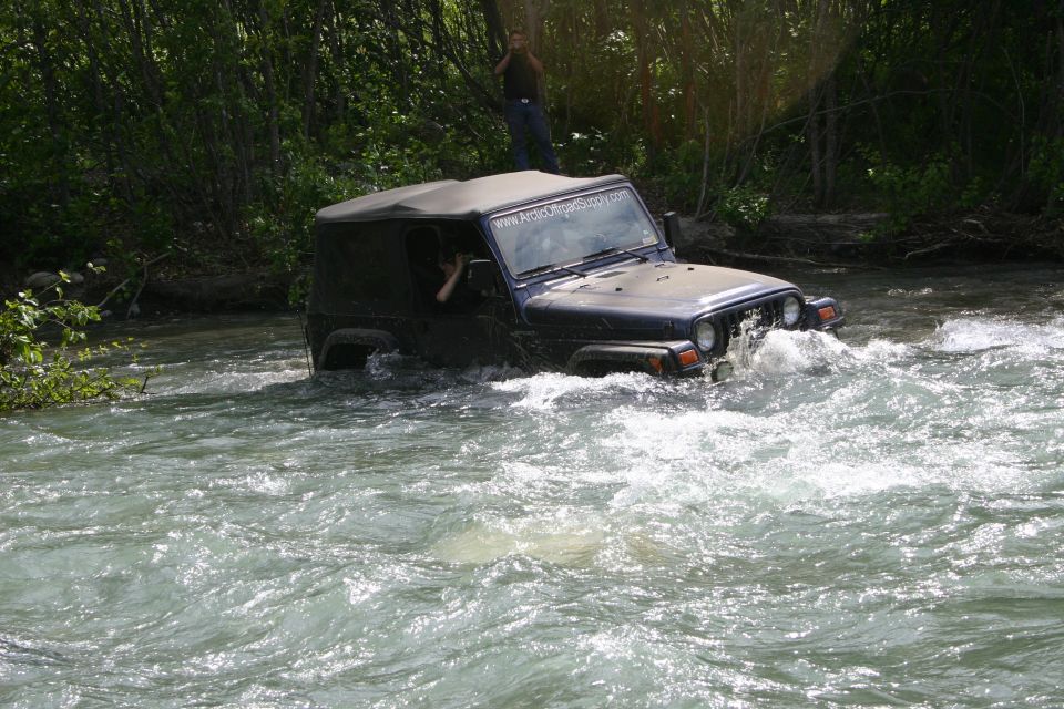 Dark blue Jeep TJ driving through a river with trees on the bank. A person stands on a rock in the background