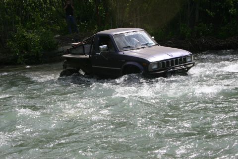 2003 Jeep TJ - A modified pickup truck navigates a rocky riverbed, with trees lining the bank.