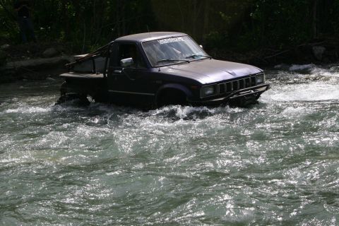 2003 Jeep TJ - A dark purple pickup truck navigates through a fast-flowing river, with trees and a person visible in the background.