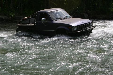 2003 Jeep TJ - A dark blue pickup truck navigates through a fast-moving river, equipped with a winch bumper for off-road challenges.