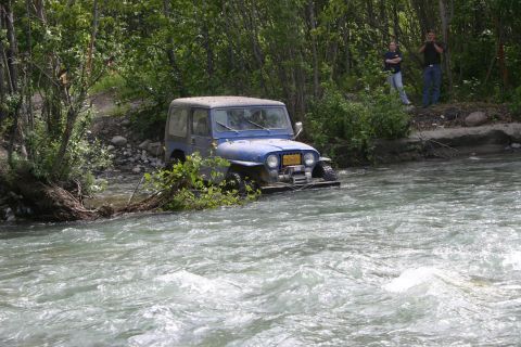 2003 Jeep TJ - A blue Jeep Wrangler navigates a rocky riverbed, with onlookers observing from the tree-lined shore.