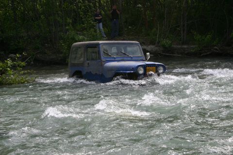 2003 Jeep TJ - A blue Jeep Wrangler navigates through a fast-moving river, with two people watching from the bank.