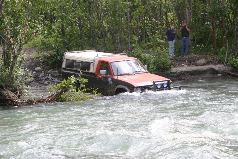 Pickup truck with a camper shell driving through a river, two people watch from the bank