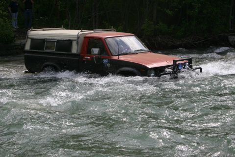 2003 Jeep TJ - A red and black pickup truck navigates a fast-flowing river, its front end submerged in the water.