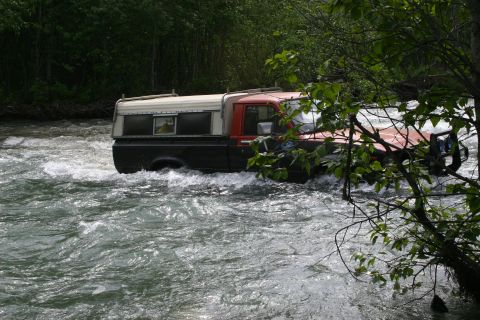 2003 Jeep TJ - A pickup truck navigates a rough river, with trees and foliage framing the scene.