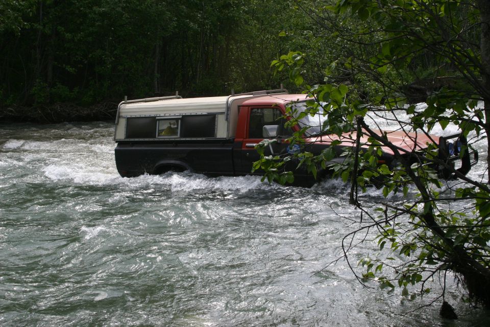 Pickup truck with a camper shell driving through a fast-moving river with trees on the bank