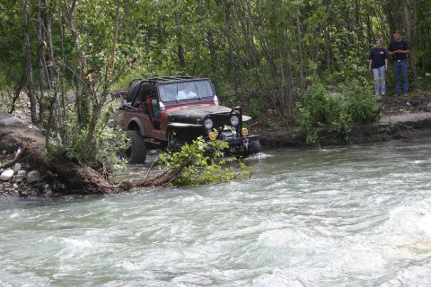 2003 Jeep TJ - A red Jeep CJ navigates a rocky riverbed surrounded by dense foliage.