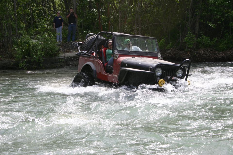 Red Jeep CJ with black fenders and roll cage driving through a river. Two people are in the front seats