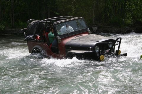 2003 Jeep TJ - A red and black Jeep Wrangler navigates through a fast-flowing river, its occupants visible inside.