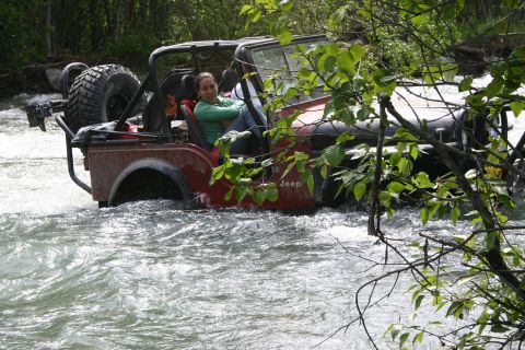 2003 Jeep TJ - A woman drives a red Jeep Wrangler through a fast-moving river, surrounded by trees.