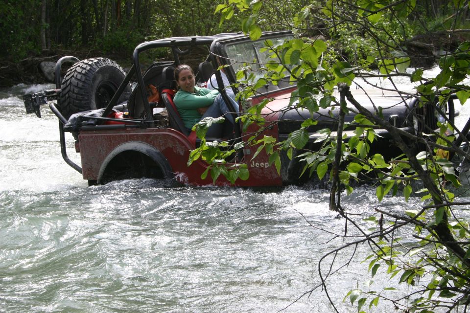 Red Jeep Wrangler driving through a river with a woman inside. The Jeep has a spare tire mounted on the back