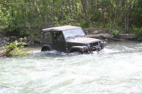 2003 Jeep TJ - A dark Jeep Wrangler TJ navigates a rocky riverbed surrounded by trees.
