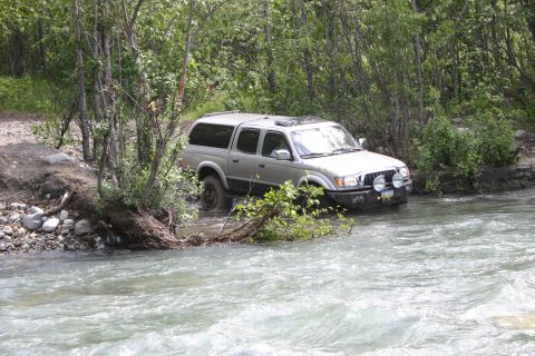 2003 Jeep TJ - A silver pickup truck navigates a rocky riverbed surrounded by lush green trees.