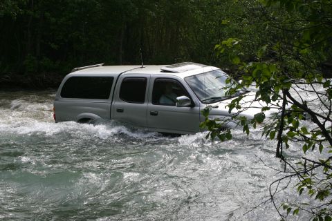 2003 Jeep TJ - A silver Toyota Tacoma navigates a rough river crossing, with trees lining the banks.