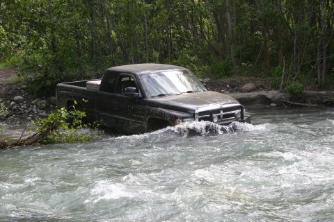 2003 Jeep TJ - A black pickup truck navigates through a fast-moving river surrounded by trees.