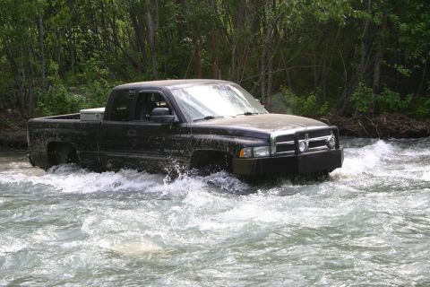 2003 Jeep TJ - A black Dodge Ram pickup truck navigates through a shallow, rocky river with trees lining the banks.