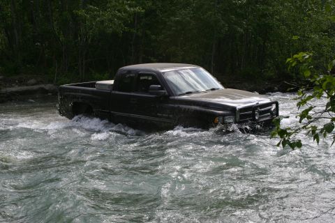 2003 Jeep TJ - Pickup truck navigates a river crossing, with trees lining the banks.
