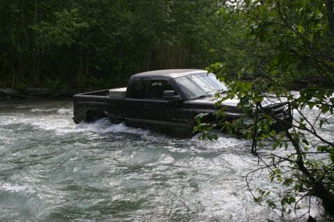 2003 Jeep TJ - A black Dodge Ram pickup truck navigates a rough river crossing surrounded by dense foliage.