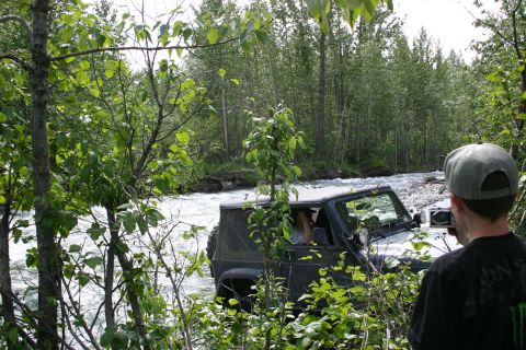 2003 Jeep TJ - A person films a Jeep Wrangler as it navigates a shallow river surrounded by dense forest.