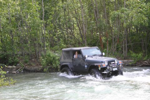 2003 Jeep TJ - A dark gray 2003 Jeep TJ navigates a river, surrounded by lush green trees.