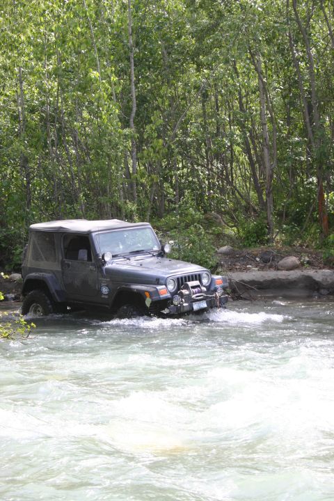 2003 Jeep TJ - A dark gray 2003 Jeep Wrangler TJ navigates through a rocky riverbed surrounded by trees.