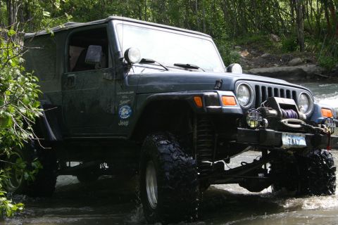2003 Jeep TJ - This Jeep Wrangler is navigating a stream, with lush green foliage and a rocky riverbed surrounding it.