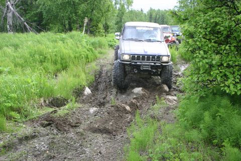2003 Jeep TJ - A blue SUV navigates a challenging muddy and rocky off-road trail with other vehicles visible behind it.