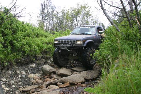2003 Jeep TJ - This modified pickup truck is driving through a challenging, rocky off-road trail surrounded by dense vegetation.