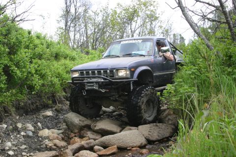 2003 Jeep TJ - A modified pickup truck navigates a challenging rocky trail surrounded by dense foliage.