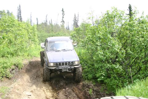 2003 Jeep TJ - A blue pickup truck navigates a challenging muddy incline on an off-road trail.