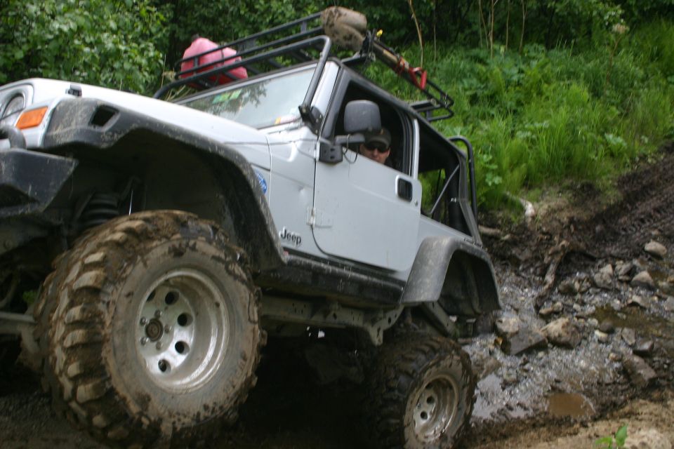 Silver 2003 Jeep TJ with large off-road tires navigating a muddy, rocky incline. Roof rack is loaded