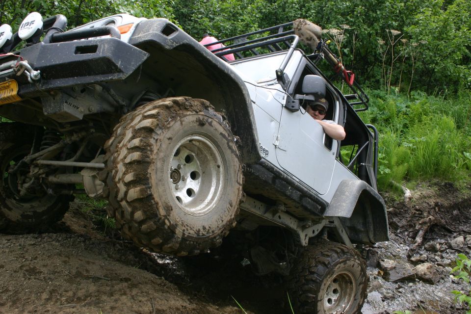 Silver Jeep TJ with off-road tires climbing a muddy incline, driver visible
