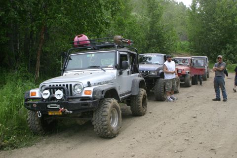 2003 Jeep TJ - A group of off-road vehicles are parked on a dusty trail surrounded by dense foliage.