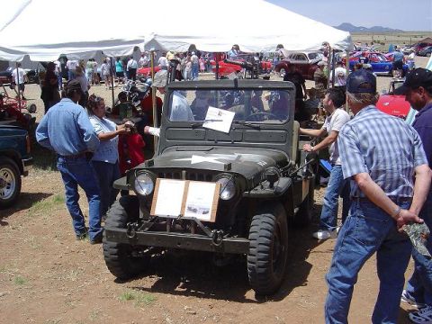 1986 Ford Bronco II - A military-style jeep is displayed at an outdoor gathering with people milling around.