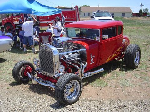 1986 Ford Bronco II - A custom red hot rod coupe with flame decals and exposed V8 engine is displayed outdoors.