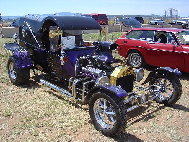 Custom hot rod with purple flames, exposed engine, and chrome exhaust pipes parked next to a red station wagon
